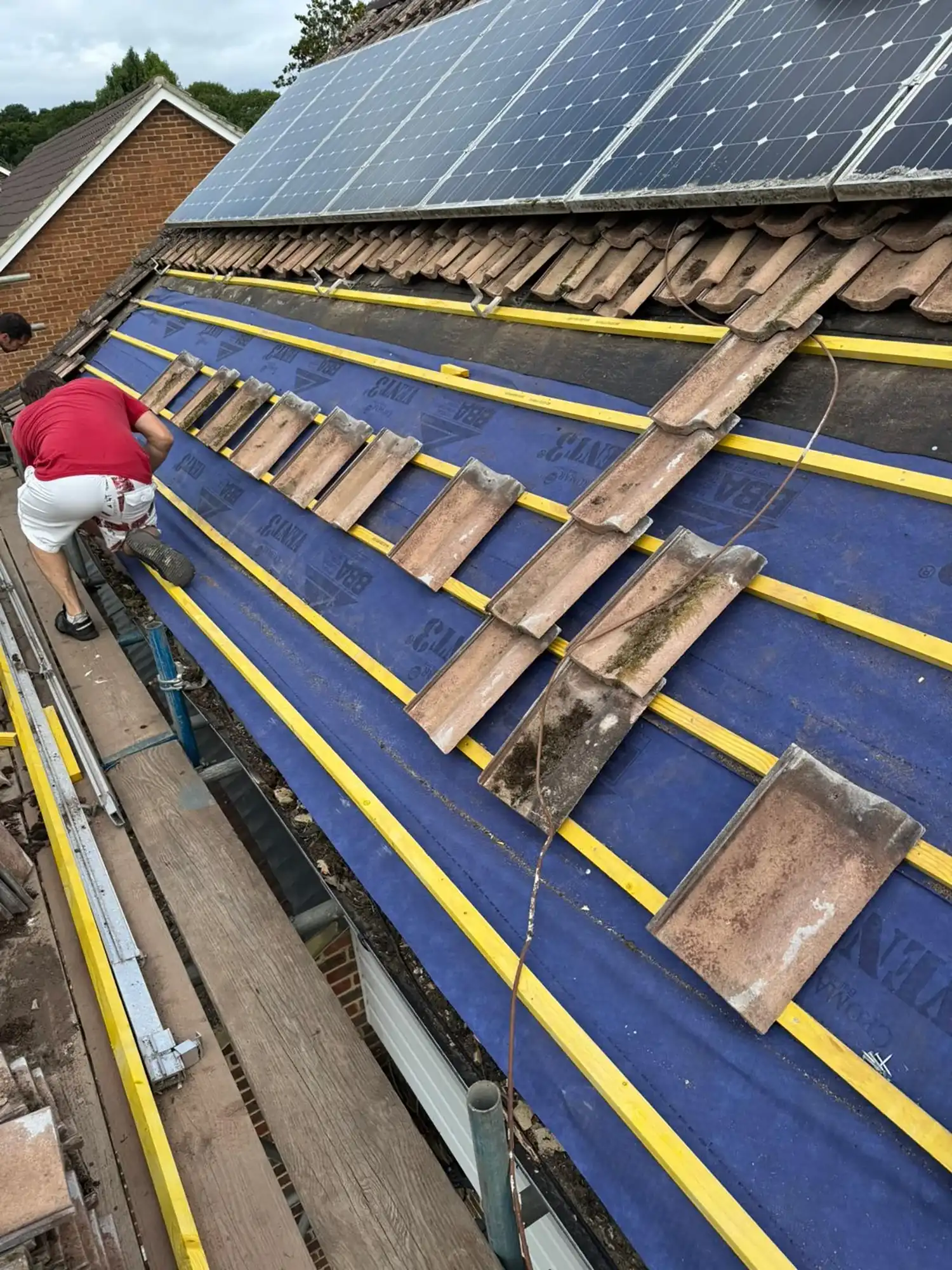 Workers installing roofing tiles on a sloped roof with solar panels on the adjacent roof section.