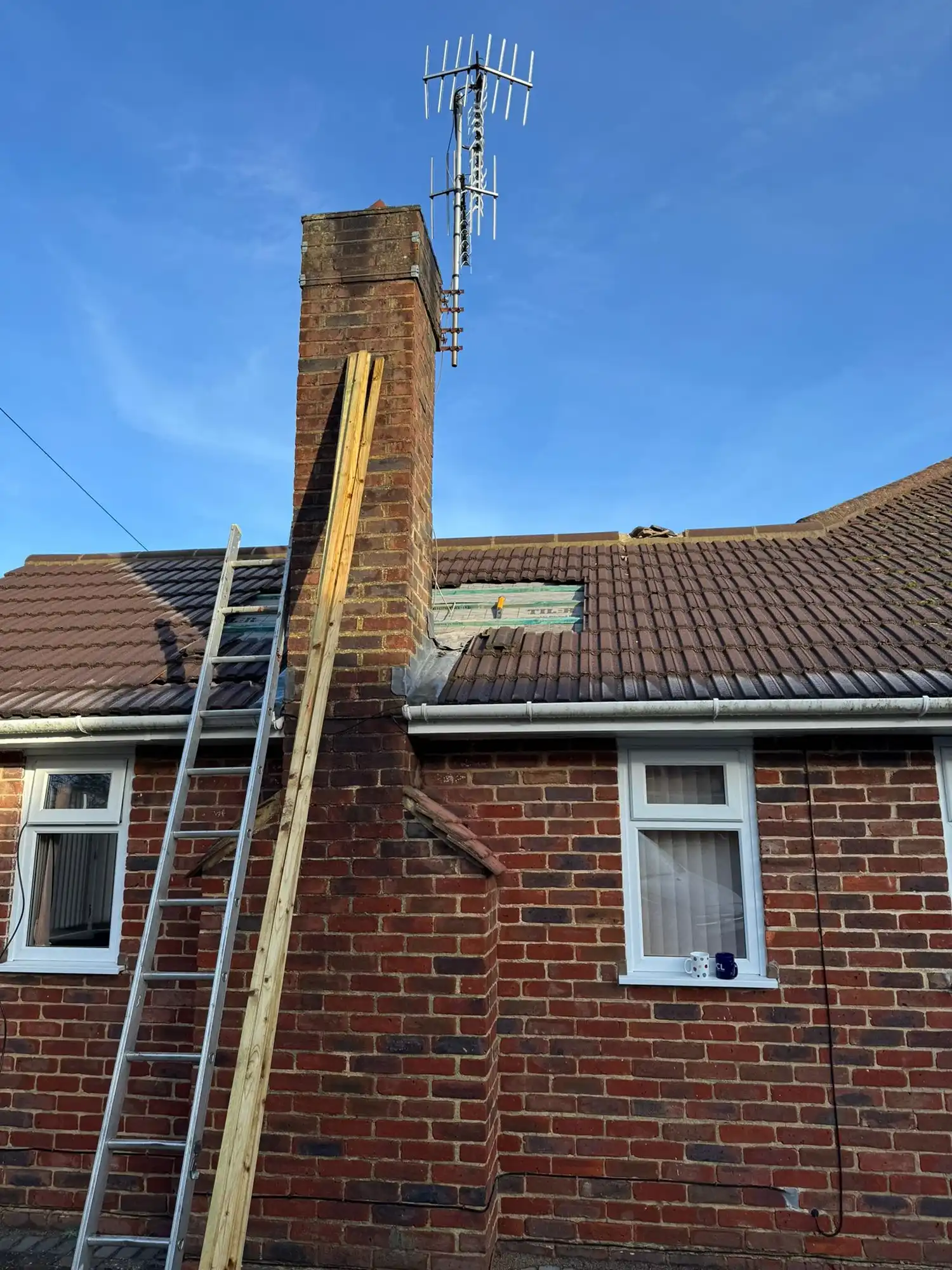 Brick house with a chimney, metal ladder and wooden planks leaning against it under a clear blue sky.