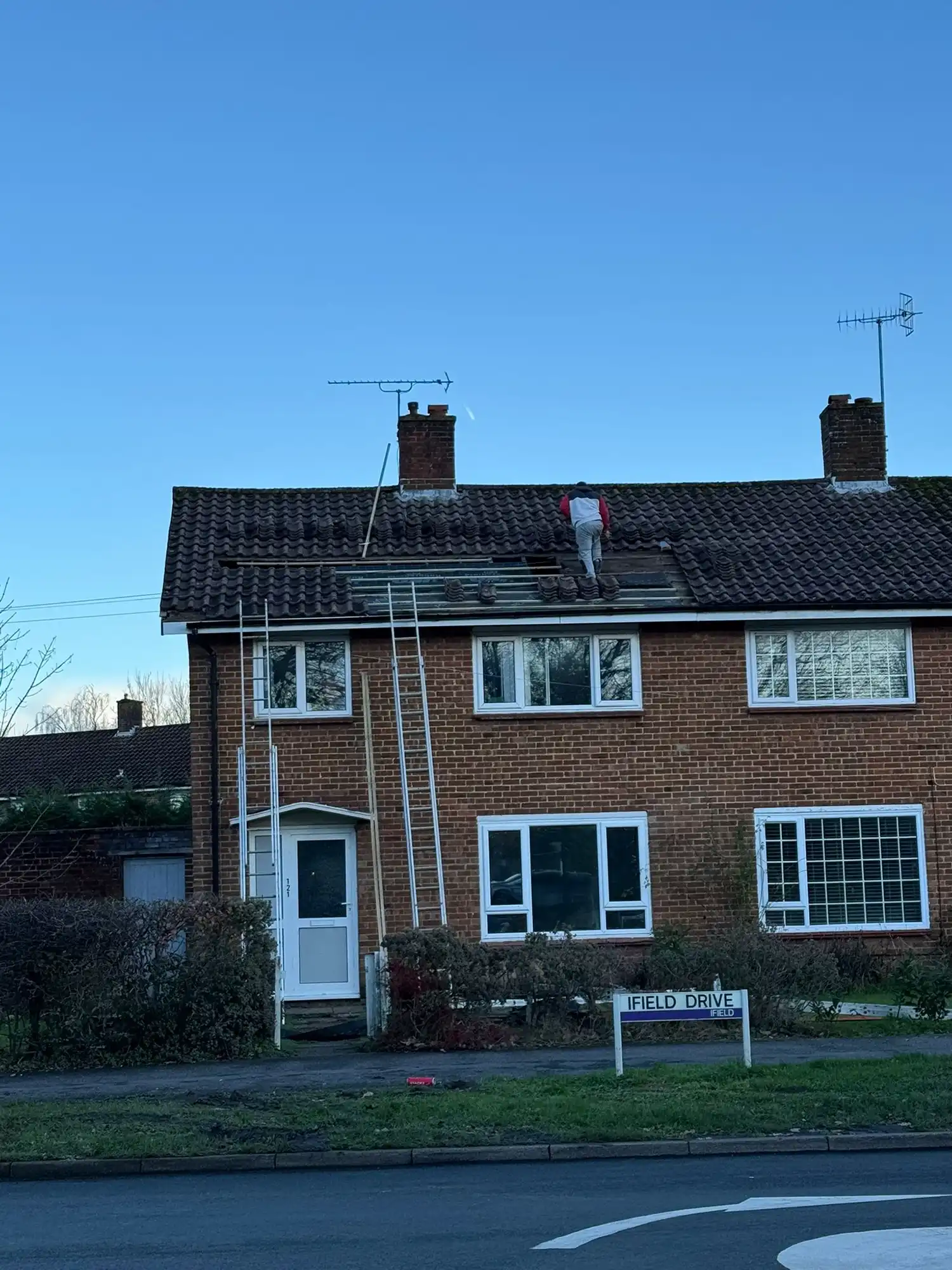 Person repairing roof tiles on a two-story brick house with two ladders against the wall.