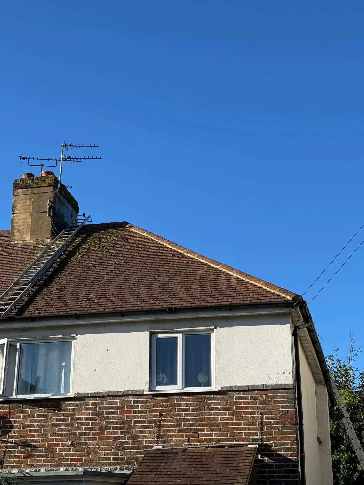Side of a brick and white house with a pitched roof, chimney, antennas, and ladders against a clear blue sky.