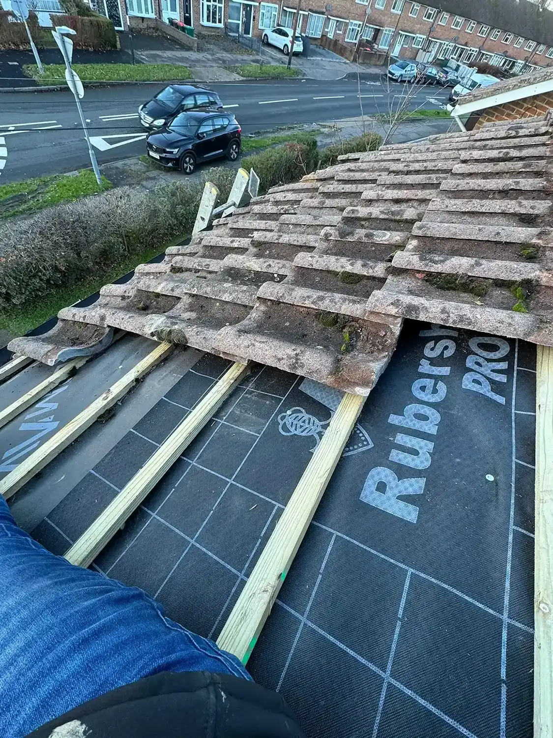 Partially removed roof tiles exposing wooden battens and roofing underlayment with the word Rubers on it.
