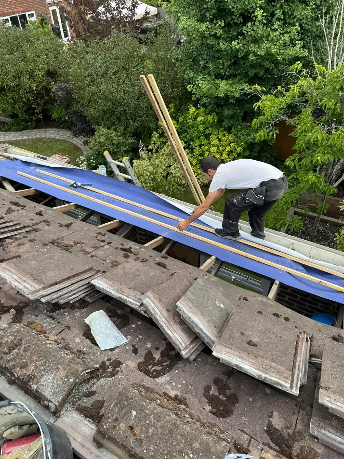 Worker measuring wooden planks on blue sheathing on a partially roofed house surrounded by trees and garden.