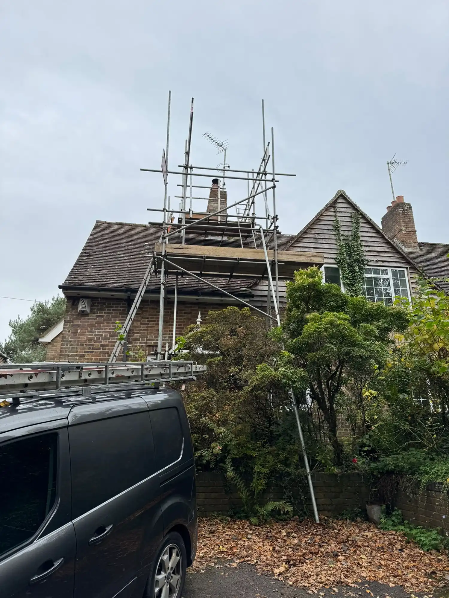 Scaffolding set up around a chimney on a brick house with a black van and leafy trees nearby.