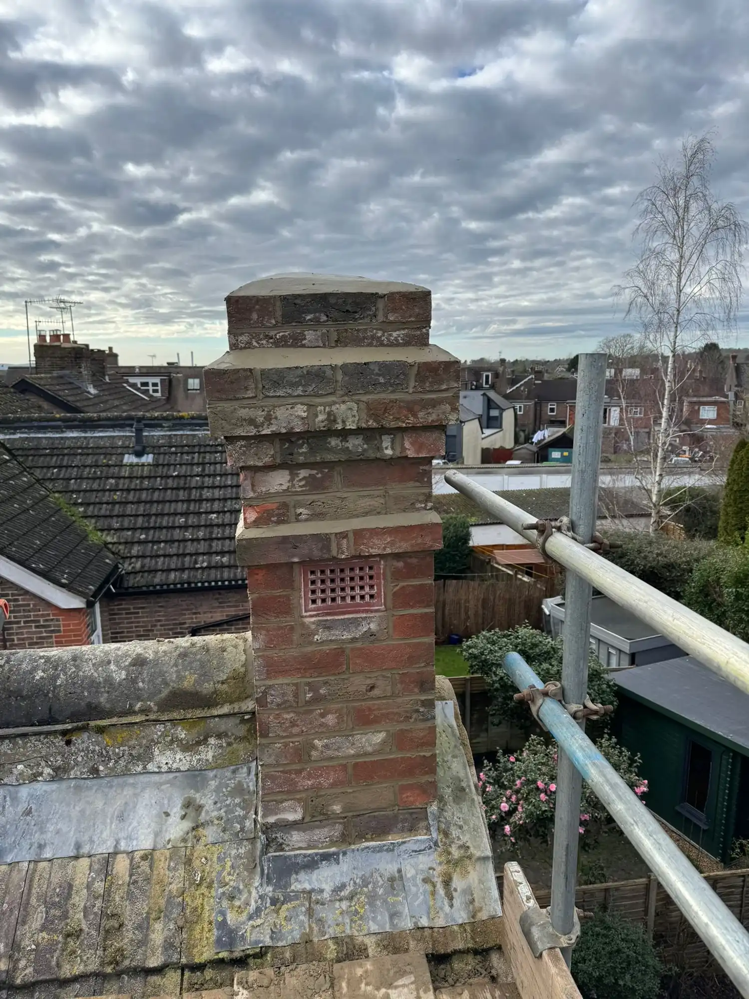 Brick chimney on a rooftop with scaffolding and residential houses under a cloudy sky.