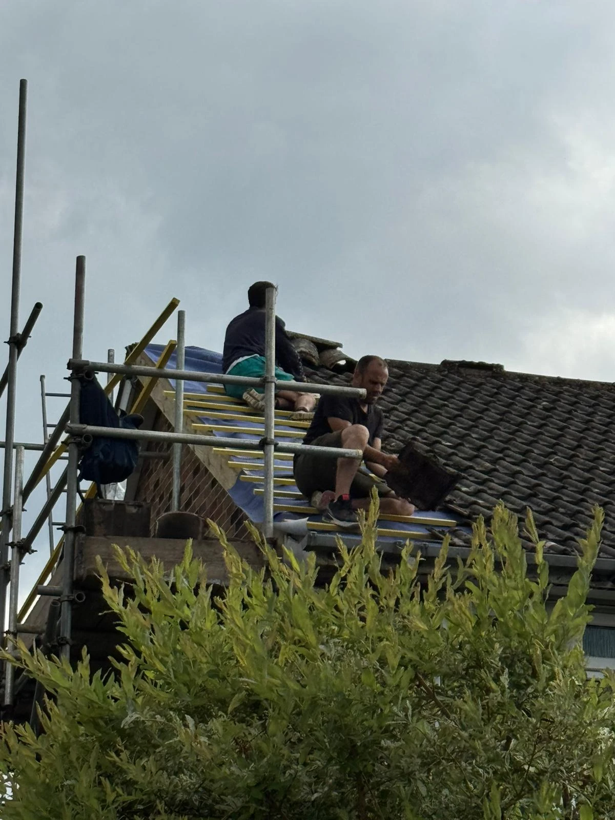 Two men working on a tiled roof with scaffolding around and greenery in front under a cloudy sky.
